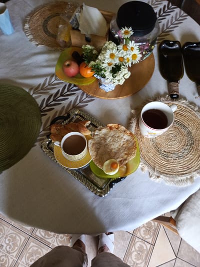 The image shows a table set for breakfast in Alexandrovskaya, Russia. On the table, there is a tray with a croissant, a cup of coffee, and a flatbread with a hard-boiled egg and a piece of fruit. There is also a mug of tea on a woven placemat, a vase of daisies, a jar, and two dark green glass bottles. The table is covered with a white tablecloth. In the foreground, a person's legs and feet are visible, wearing white slippers and brown pants. The floor is tiled.