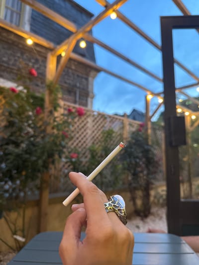 An individual's hand, adorned with a unique ring, holds a lit cigarette. The cigarette is positioned in the foreground, angled to the left, with its glowing tip illuminated. The hand itself is in focus, revealing details of the skin and the intricate design of the ring on the ring finger. The background is softly blurred, suggesting an outdoor setting. There are visible elements of a pergola with string lights, a wooden lattice fence, lush green foliage with pink roses, and the facade of a building. The sky above is a dusky blue, indicating it is either dusk or dawn. The overall scene conveys a moment of quiet contemplation or relaxation in a garden-like environment. The location is identified as Veules-les-Roses, France.