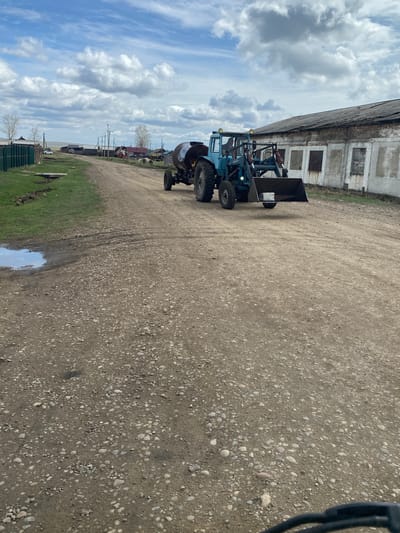 The scene depicts a rural landscape near what could be the city of Kapsal, Russia, though specific landmarks confirming the exact location are not apparent in the image. A light blue tractor with a front-end loader sits on a gravel road. Adjacent to the road is a weathered, possibly abandoned building with boarded-up windows. Beyond the road and tractor, there is a grassy area with puddles, possibly from recent rainfall. In the background, there are signs of human habitation including houses and utility poles. The sky is partly cloudy. The tractor appears to be stationary. There are no visible people or actions occurring beyond the presence of the tractor on the road.