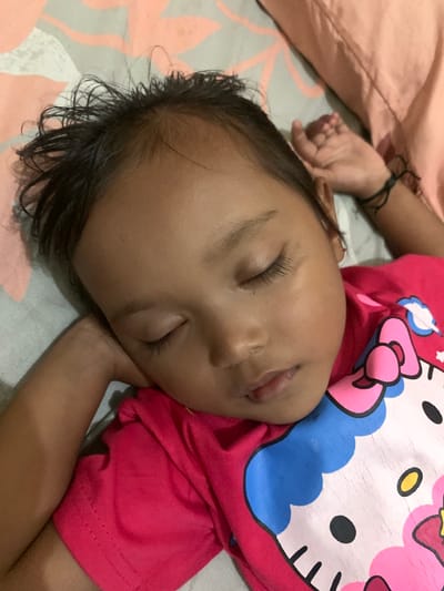 An image captured in Sukorejo, Indonesia, depicts a young child of indeterminate gender, likely of East or Southeast Asian ethnicity, sleeping. The child is positioned on their left side, facing slightly right, with eyes closed. Dark, somewhat tousled hair is visible on the child's head. The child wears a short-sleeved, bright pink or fuchsia t-shirt featuring a worn "Hello Kitty" graphic on the chest, characterized by visible cracking in the print. The child's left arm is bent, with the hand resting near the head. The right hand, visible in the upper right portion of the frame, rests on a peach-colored pillow or folded fabric and has a thin black string tied around the wrist. The child is lying on a light grey fitted sheet adorned with a subtle pattern of light peach or pink abstract shapes. The overall lighting suggests an indoor setting.