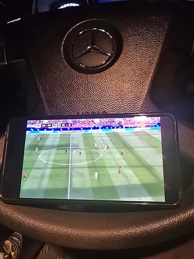 A mobile phone is placed on the steering wheel of a Mercedes-Benz. The phone displays a soccer game with a score of 1-1. The game time is 71:46. The steering wheel is dark leather with the Mercedes-Benz logo in the center. In the background, a dashboard with illuminated lights is visible. The image is taken from a low angle, looking up at the steering wheel and phone. The environment appears to be the interior of a car.