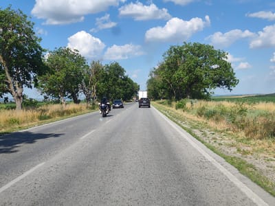 A forward-facing, eye-level shot captures a rural road in Totleben, Bulgaria, on a sunny day with scattered clouds. The asphalt road is divided by a white line, with a shoulder of dry grass and scattered green bushes lining both sides. A row of mature trees with lush green canopies flanks the road, creating a shaded tunnel effect in some areas. In the foreground, the road is empty, but further ahead, a motorcycle with a rider in black is traveling in the left lane. Behind the motorcycle, a dark sedan is also in the left lane. In the right lane, a dark SUV is visible, and further down the road, a white truck is traveling in the distance. The background reveals a vast, open landscape of fields stretching to the horizon, under a bright blue sky dotted with white cumulus clouds.