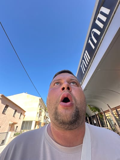 A wide-angle, low-angle, outdoor, selfie-style photograph captures a man with his mouth agape, looking upwards. The man, with light skin, a beard, and wearing a light-colored t-shirt, occupies the foreground. To the upper right of the frame, a dark sign with white lettering, reading "TAM TAM", is visible against a clear blue sky. In the background, to the left, are two buildings with light-colored facades and tiled roofs. The architecture suggests a European setting, consistent with the location of Platja D'aro, Spain. A thin black wire stretches diagonally across the sky from the upper left to the middle right of the image. The lighting indicates it is a sunny day.