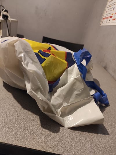 A crumpled white plastic shopping bag with blue and yellow branding, possibly from Lidl, lies on a speckled grey countertop. The bag appears to be empty and is partially collapsed, with its handles made of twisted blue plastic. The background consists of a grey wall and a dark surface that may be a chair or table. To the left, a power strip with cords is visible, plugged into an outlet on a white box. A poster with text and icons is affixed to the wall in the upper right corner. The scene suggests an indoor setting, likely a home or commercial establishment, with no people present. The lighting is dim, casting shadows on the countertop. The image was taken in Chaponost, France.