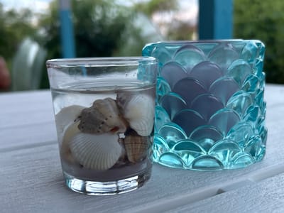 The image shows a close-up of two decorative items on a white wooden surface. On the left is a clear glass filled with water and seashells. The seashells are various types and sizes, with some being white and others a sandy brown color. The water level is high, nearly reaching the rim of the glass. To the right of the glass is a teal-colored glass candle holder with a textured, scalloped design. The candle holder appears to be empty or contain a candle that is not visible. The background is blurred, showing greenery and what appears to be a blue structure, possibly part of an outdoor setting. The lighting suggests it is daytime. The location is not specified in the image.