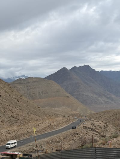 The image depicts a mountainous landscape near Chumkar, India, under an overcast sky. A paved road winds through the rugged, rocky terrain, with several vehicles visible: a white van is parked on the left side of the road, a black SUV is driving on the road, and a white car is further down the road. The mountains are barren and characterized by layered rock formations. In the foreground, there are remnants of structures, including corrugated metal fencing and what appears to be a small building with a red-and-white roof. A yellow flag on a pole is also visible near the road. The overall scene is one of a remote, arid, and mountainous region.