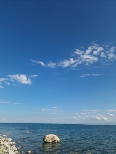 This media file depicts a seascape with a clear blue sky and a calm body of water. The sky is largely a deep blue with scattered white, wispy clouds. The horizon line is distinct, separating the sky from the water.
The water is a deep blue-green and appears to be a lake or sea. Gentle ripples suggest a light breeze. On the right side of the image, the shoreline is visible, characterized by a scattering of light-colored rocks and stones, some submerged in the water. A prominent, large, light-colored boulder rests partially in the water near the shore, casting a subtle reflection. The water's edge is rocky and appears to be a natural, undeveloped area.
The image conveys a sense of tranquility and natural beauty. The bright sunlight suggests it is daytime, likely late morning or early afternoon, given the clear, vibrant sky. There is no visible text in the image. The scene is likely a natural coastal or lakeside environment. Given the location context of Sevan, Armenia, this could be a view of Lake Sevan.