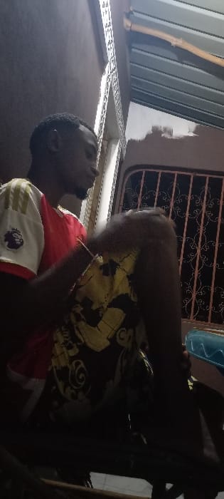 A young adult male, dark-skinned with short dark hair and a beard, is depicted seated in profile facing right. He wears a red and white short-sleeved t-shirt with a "Premier League" patch and the number "7" on the sleeve, and black shorts with yellow patterns. A light-colored bracelet is visible on his left wrist. His left knee is bent and raised. The background features a plain dark wall to the left, a white vertical decorative trim, and an ornate pinkish-brown metal-grilled window or door to the right. Above the window, a corrugated metal roof section is visible, with a patch of lighter-colored paint on the wall below it. The scene is dimly lit, suggesting an evening or interior setting in Kinondo ward, Kenya.