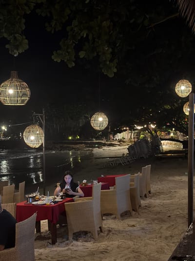 This image captures a nighttime outdoor dining scene on a sandy beach in Ao Phai, Thailand. The primary focus is a female individual seated at a table on the sand in the foreground-midground. She possesses long dark hair and wears a dark top with light-colored shoulder detailing. She is observed looking down at a white smartphone held in both hands, with her mouth slightly agape. To her immediate left, the back of a male individual's head and shoulder are visible, suggesting another diner at the same table.
The dining table is covered with a red tablecloth and surrounded by light-colored woven rattan-style chairs. On the table are several items, including empty or partially consumed white plates with food residue, glasses containing orange-colored beverages, cutlery, and a metal pot on a stand.
The environment is dimly lit by numerous spherical lanterns crafted from woven material, suspended at various heights from tree branches and a visible wooden pole. These lanterns emit a warm, yellow light, reflecting on the wet sand near the water's edge. Beyond the dining area, the ocean or sea is visible on the left, displaying dark water with reflections of ambient lights and small breaking waves. The sandy beach extends into the background, where clusters of white stacked beach chairs and loungers are discernible under the dark foliage of trees. Further in the background, additional indistinct lighting suggests other resort structures or dining areas along the coastline. The sky is dark, indicating night.