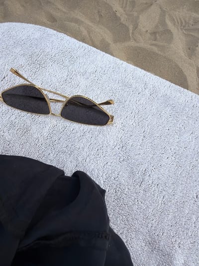 The image shows a close-up, high-angle shot of a white towel on a sandy beach. The towel has a plush texture and is partially covered by a black garment in the lower left corner. Resting on the towel are a pair of gold-rimmed sunglasses with dark lenses. The background consists of rippled sand, indicating a beach environment. The scene is devoid of any people or visible human activity, suggesting a tranquil moment. This scene is located in the city of Olympiada, Greece.