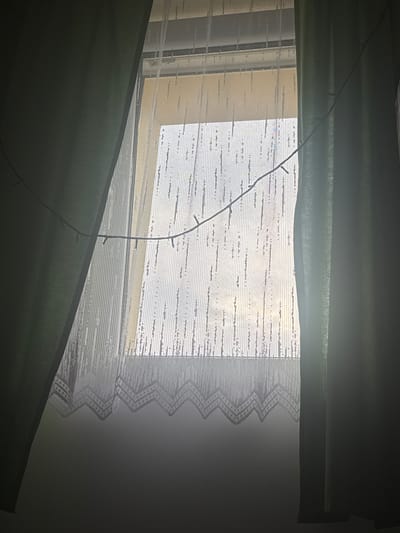 This image captures a dimly lit interior scene, likely a bedroom, with a window as the central focus. The window is covered by layered curtains. The innermost layer appears to be a sheer, white lace curtain with a decorative zig-zag pattern along its edge. Behind this, there are blinds or a roller shade, visible as horizontal bands of fabric. The outermost layer consists of thick, dark green curtains, pulled partially closed.
A string of small, possibly LED, lights is draped across the window, extending from the top left towards the bottom center of the image. These lights are not illuminated. The overall lighting suggests it might be nighttime or a very overcast day, as the light filtering through the window is soft and diffused. The scene is still and quiet, with no people visible. The focus is on the textures of the fabrics and the interplay of light and shadow. The location context provided is Marki, Poland, but there are no specific visual cues within the image to confirm this.