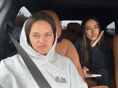 Two women are in a car, appearing to take a selfie. One woman, on the left, has long brown hair, is wearing a dark-colored jacket, and is holding a smartphone. The other woman, on the right, is wearing a grey hooded sweatshirt with the words "The Club is Closed" printed on it, and is wearing a seatbelt. The car's interior is brown leather. The location cannot be determined to be Verin Ptghni, Armenia from the given information.