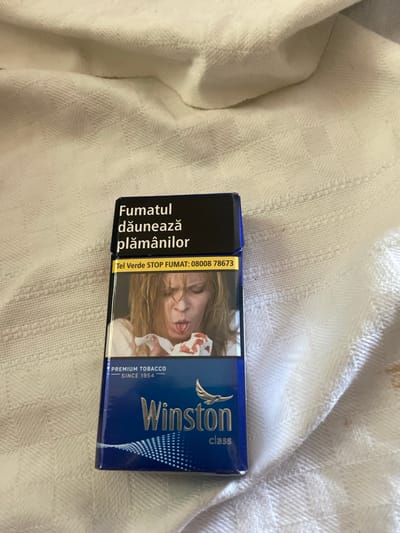 An open pack of Winston cigarettes is positioned on a white, textured fabric. The pack is predominantly blue with a black top panel. The black panel displays text in white and yellow, with the prominent Romanian phrase "Fumatul dăunează plămânilor" (Smoking harms the lungs) in white. Below this, a yellow band contains the text "Tel Verde STOP FUMAT: 08008 7673".
The front of the pack features a graphic image of a woman with blonde hair, her face contorted as if coughing, holding a white tissue stained with blood in front of her mouth. Below this image, in white text, are the words "PREMIUM TOBACCO SINCE 1954". The bottom portion of the pack is blue and displays the Winston logo, a bird in flight, and the word "class" in white text.
The fabric beneath the cigarette pack appears to be a blanket or tablecloth with a subtle checkered pattern. The lighting suggests an indoor environment. The overall impression is one of the packaging of a tobacco product, with a prominent health warning. There are no people present in the image other than the depicted graphic. The location is Unknown, Unknown.