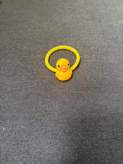 A small, yellow rubber duck charm is attached to a yellow elastic hair tie, which is looped on a dark gray, textured surface. The duck is positioned in the center of the hair tie, facing forward. Its features, including its beak and eyes, are clearly visible. The background is a plain fabric with a subtle woven pattern. The image is taken from a top-down perspective. This scene is not located in Koshkino, Russia.