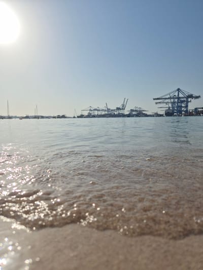 The image depicts a low-angle view of a sandy beach with gentle waves lapping at the shore. The water is clear and reflects the bright sunlight, creating shimmering highlights. In the background, a port facility is visible, characterized by several large cranes silhouetted against a clear blue sky. Sailboats and other vessels are also present in the distance, suggesting a coastal or harbor area. The overall scene is one of a sunny day at the seaside, with a juxtaposition of natural elements and industrial infrastructure. The location is identified as Birżebbuġa, Malta, a town known for its significant maritime activities and port facilities.