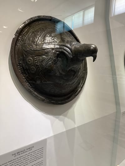 The image shows a close-up view of a large, dark bronze shield with intricate carvings, displayed in a museum case. The shield is convex and has a prominent bird-like projection extending from its center. The surface of the shield is decorated with repeating patterns, including circular motifs and what appear to be stylized animal figures. The bronze exhibits signs of age and wear, with some cracks and imperfections visible.

The shield is displayed against a plain white background, which is slightly obscured by the glass of the display case, causing reflections. A label in the lower left corner of the image reads "THE BRONZE SHIELDS OF THE IDAETAN CAVE". The text describes the shields as being from the Idaetan cave, made of bronze, and used as lavish votive offerings. It mentions their ornamental decoration, hammered and incised designs, and inclusion of frontal animal motifs, as well as human figures. The text also hints at the geographical location by referencing the Idaetan cave.

While the image is taken within a museum, the specific location in the city of Heraklion, Greece is not explicitly stated. However, given the label mentioning the "Idaetan cave," it is highly probable that this artifact is displayed at the Archaeological Museum of Heraklion, which houses many important Minoan and Mycenaean artifacts, including those from Mount Ida. The presence of a label and the museum display setting suggest that this is a historical artifact being exhibited to the public. No people are visible in the image, and no actions or interactions are occurring. The environment is that of a museum exhibit.