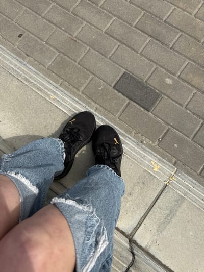 A low-angle, overhead shot captures a person's lower body, focusing on their legs and feet in an outdoor setting in Ahronomichne, Ukraine. The individual is wearing light blue denim shorts with heavily frayed and distressed edges, revealing bare skin on their legs. On their feet are black sneakers, each featuring a prominent gold Puma logo on the outer side. The person appears to be standing or sitting on a concrete or stone curb. The ground beneath their feet is paved with rectangular grey concrete blocks, laid in a diagonal pattern, typical of a sidewalk or pedestrian area. A distinct concrete curb or step runs diagonally across the image, separating the paved area from a lighter, smoother concrete surface. Some yellow material, possibly sealant or paint, is visible in the joints and cracks along the curb. The bright, direct lighting and clear shadows indicate it is daytime, likely sunny.