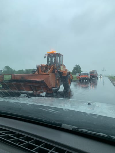 An orange front-end loader is stopped on a wet road. The loader has a large bucket in front and an orange light bar on top. In the background, there are several vehicles stopped on the side of the road, including a white and orange van and a red truck. The road appears to be a rural road with trees and power lines visible in the distance. The sky is overcast and it appears to be raining. The scene is located in the city of Kurapolski Selski Savet, Belarus.