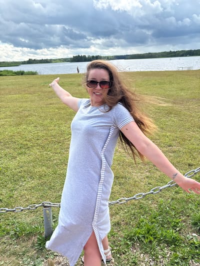 A woman with long, brown hair and sunglasses stands in a grassy area near a body of water. She is wearing a light gray dress with white stripes down the side. Her arms are outstretched, and she is smiling at the camera. The background shows a lake, trees, and a cloudy sky. There are also two people visible in the distance near the water. A metal chain fence is in the foreground. The location is identified as Mazalawski Sel'ski Savelet, Belarus.