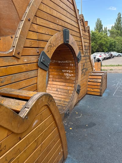This is a close-up, slightly angled, outdoor photograph of a wooden playground structure. The structure appears to be made of large wooden planks and is shaped in a way that suggests a ship or a similar vessel. It is adorned with various graffiti and handwritten inscriptions, some of which are legible.

In the background, through a circular opening in the wooden structure, a few cars are visible parked on a paved surface, along with trees and a clear blue sky, indicating it's daytime and likely sunny. The ground in front of the structure is dark, possibly asphalt, with some chalk markings, suggesting a recreational area.

Visible text in the image includes names and possibly dates or short phrases in Cyrillic script. Examples include "АРТЕМ+", "НИКИТА + УЛЬЯНА =", "КАТЯ ЗБУГИ", and "Ягона из зв". There are also some hearts and other symbols drawn on the wood. The overall impression is of a well-used, outdoor play space in a Russian setting.
