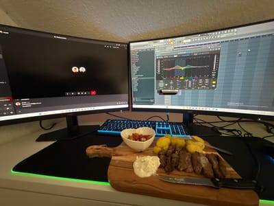 The scene depicts an indoor desk setup in Vellmar, Germany. Two black widescreen computer monitors are positioned side-by-side on a white desk. The left monitor displays a dark video conferencing interface with two user avatars visible and a chat panel on the left. The right monitor displays a digital audio workstation (FL Studio) with an audio plugin (identified as "AUTOTUNE Vocal B") showing an equalizer graph and various controls.
A black mechanical keyboard with blue backlighting is situated in front of the monitors. A large black mousepad with a green LED illuminated edge covers the central desk area. Resting on the mousepad is a light brown wooden serving board. On this board, a meal is arranged: approximately seven pieces of seared meat, a pile of cooked yellow potatoes, a dollop of white creamy sauce, and a small white bowl containing a chopped red and white vegetable mixture (salsa). A black-handled serrated steak knife is positioned on the board next to the meat, and a silver fork is partially visible near the potatoes. Cables are visible behind the keyboard and monitors. No individuals are physically present in the image.