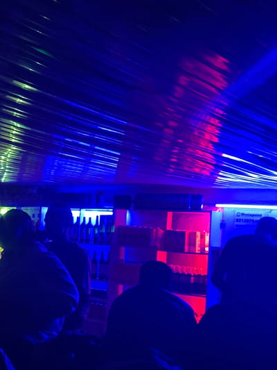 The image depicts an indoor scene, possibly a bar or club, illuminated by vibrant blue and red lights. In the foreground, silhouettes of several individuals are visible, facing away from the camera. The individuals appear to be standing or sitting. The background reveals shelves stocked with bottles, some of which are illuminated by the colored lighting. A sign with text, including "Moniepoint" and a numerical code, is partially visible on a white surface in the mid-ground. The ceiling is covered with what appears to be corrugated material, reflecting the light and creating a striped effect. The overall atmosphere is dark and dimly lit, with the colored lights creating a dramatic effect. The location is identified as Wumat, Nigeria.