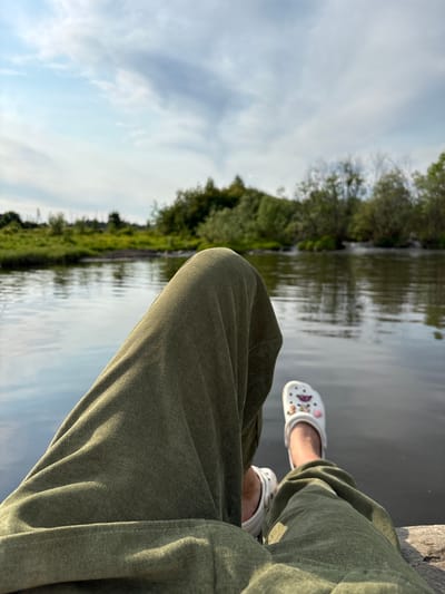 The image shows a close-up perspective of a person's legs and feet, clad in olive green pants and white Crocs, resting on a stone ledge by a body of water. The water's surface reflects the sky and the surrounding lush green trees and vegetation, creating a serene natural landscape. The sky is partly cloudy, with patches of blue visible through the white clouds. The overall scene suggests a moment of relaxation in an outdoor setting, possibly near the city of Nevon, Russia, given the natural environment.