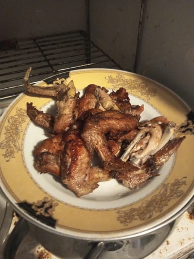 The image displays a white ceramic plate, featuring a broad yellow decorative band with light brown floral patterns along its rim and distinct black ornate segments. The plate is laden with numerous pieces of cooked poultry, primarily consisting of browned chicken wings and other small chicken cuts, exhibiting varied textures from crispy skin to exposed bone and lighter flesh. This culinary scene is located in B. Nam Ngao, Laos. The plate rests on an indistinct surface, with a metallic object partially visible below its left edge. In the background, an interior chamber, likely an oven, is visible, containing a metal wire rack across the upper left and showing signs of use and minor discoloration on its light-colored walls. No individuals are present in the frame; the scene captures a still life of prepared food within a kitchen environment.