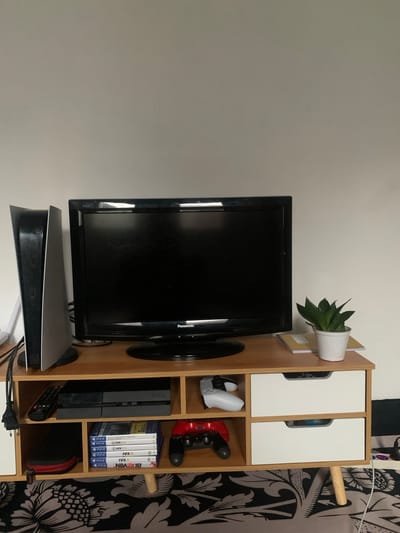 A television set, gaming console, and related accessories sit on a shelf, appearing to be indoors. A flat screen television is positioned towards the left of the frame, with a Panasonic logo visible on the upper right corner of the screen frame. Underneath the television, a Playstation 5 console rests. To the right of the television, the shelf contains gaming accessories: two Playstation controllers, one white and one red, a Playstation 4 console, several FIFA games (15, 16, 17, and 18), and NBA2K18. In the top left of the shelf, a potted succulent is placed. The shelf unit itself is made of light wood with two white cabinet doors at the top. The background includes a white wall to the left and a patterned curtain on the right. The location is not specified or identifiable in the image.
