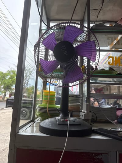 In Meunasahmanyet, Indonesia, a close-up shot features a black electric fan with purple blades positioned on a shelf within a glass display case. The fan is in focus, its metallic grate and vibrant blades clearly visible. Behind the fan, stacks of colorful plates, some green, yellow, and orange, are arranged on another shelf. The display case reveals a glimpse of the outdoors through its glass panels, showing trees and a cloudy sky, suggesting a daytime setting. A power cord from the fan is visible, along with a white electrical adapter plugged into a nearby outlet. To the right of the fan, a dark-colored smartphone lies on the shelf, partially obscuring a round, clear lid. The overall scene appears to be an indoor space adjacent to a street or outdoor area, possibly a shop or eatery, given the display of plates.