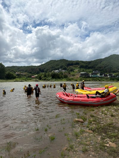 A group of people, most wearing yellow life vests and some with helmets, are in a shallow river, some standing and others wading. Two red inflatable rafts are positioned in the foreground, one partially in the water and one on the rocky bank. A yellow inflatable raft is further out in the water. In the background, a lush, tree-covered hillside rises, with buildings scattered on its slopes and a road visible. The sky is overcast with white and gray clouds. This scene is located in Seo-myeon, South Korea.