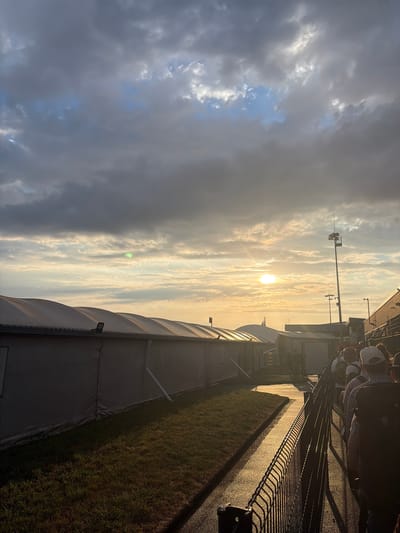 The image shows a queue of people lined up outdoors, with a row of white tents or temporary structures to their left. The sun is setting or rising, casting a warm, golden light across the scene and creating long shadows. The sky is partly cloudy, with dramatic clouds in shades of gray and white, and patches of blue visible. In the distance, there are buildings and what appear to be lighting towers. The ground is a mix of asphalt and grass. The overall impression is of an event or gathering taking place outdoors at dusk or dawn. The location is identified as the city of Tillé, France.