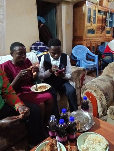 A group of people is seated on couches, enjoying a meal. Plates of food, including rice and meat, are placed on a wooden table alongside several bottles of dark liquid with blue caps. One man, dressed in a vest and white shirt, is using his phone while others are eating. A large wooden cabinet stands in the background, and a blue plastic chair is also visible. The scene suggests a casual gathering or celebration.