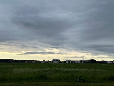 The image depicts a cloudy sky over a green field with several modern houses visible in the background. The scene is likely located in the city of Harkujärve, Estonia, given the architectural style of the buildings. The sky is overcast with a mix of dark grey and lighter, brighter clouds, suggesting either dawn or dusk. A hint of yellow light breaks through the clouds on the horizon. The field in the foreground is lush and green, dotted with small white wildflowers. The houses are a mix of dark and light structures, many with flat roofs and large windows, typical of contemporary residential architecture. There are no people or animals visible in the image, and no specific actions are taking place. The environment appears to be a suburban or developing rural area.