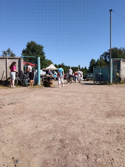 The image shows an outdoor scene with a clear blue sky and a dirt ground. In the foreground, the dirt is scattered with small rocks and debris. In the middle ground, a group of people are present, some of whom are near bicycles. There are also several parked cars and a tent in the background. The scene appears to be a market or gathering, possibly a flea market, given the presence of stalls and people browsing. The environment is rural or semi-rural, with trees visible behind the structures. The overall impression is of a casual daytime event under bright sunlight. It is possible that this scene is located in the city of Irshansk, Ukraine, as indicated in the prompt, however, there are no specific visual cues within the image to confirm this location.