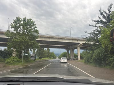 The image depicts a wet, overcast highway scene in Samtredia, Georgia. A white Toyota Prius is driving on the right lane of a two-lane road under a large concrete overpass. To the left of the road, a truck is parked on the shoulder, and a traffic sign with two circular plates is visible. Dense green trees and foliage line the sides of the road and the overpass supports. The road surface reflects the grey sky, indicating recent rain. The overpass itself is made of concrete with metal railings along its edges, and it is supported by multiple concrete pillars. In the distance, the road curves slightly and appears to lead towards rolling hills. The overall atmosphere is somber due to the cloudy weather.