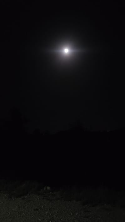 A low-angle, night shot shows a bright moon in the dark sky, appearing in the left portion of the frame. The right portion of the frame shows a dark area, with a gravelly or rocky surface in the extreme lower-right. The location is identified as Ziarat, Pakistan. There are faint lights in the distance in the upper center. No people, specific objects, actions, or interactions are visible.