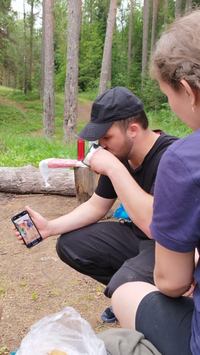 In a forest setting, a man wearing a black baseball cap and black t-shirt is crouched down, drinking from a metal mug. He holds a smartphone in his left hand, displaying a photograph of people. Beside him, a woman with her back to the camera, wearing a purple t-shirt and black shorts, is also crouched. In the foreground, a plastic bag with some items inside is visible, along with a log and scattered dry leaves and grass on the ground. The background features tall trees and a path winding through the forest, suggesting a location likely in or around Orekhovo, Russia.