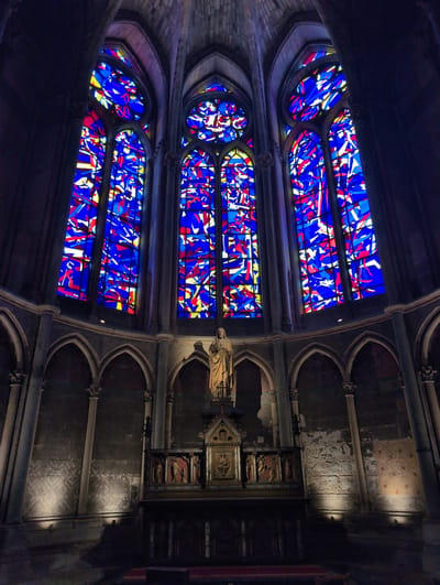 The image shows a view inside a large, Gothic-style building, likely a church or cathedral, with tall, arched windows filled with vibrant stained glass. The stained glass features abstract patterns in shades of blue, red, and yellow, with sunlight filtering through them, casting colored light onto the interior. In the foreground, below the stained glass, is an altar with a statue of a person, possibly a religious figure, standing at the top. The altar is elaborately decorated with carved panels and has a dark, wooden base. The surrounding architecture includes stone columns, arches, and textured walls, some of which are illuminated by spotlights. The overall impression is one of grandeur and historical significance. The location is identified as Urvillers, France.