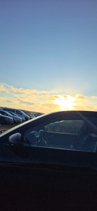 The image depicts a parking lot at sunset, with rows of cars visible under a sky filled with clouds illuminated by the setting sun. The foreground is dominated by the interior of a dark car, showing a steering wheel and part of a seat, viewed through the passenger side window. In the background, a variety of vehicles, including vans and sedans, are parked in neat rows. The sky is a gradient of blue, with streaks of orange and yellow light from the setting sun breaking through the clouds. The scene is likely set in a large open area, possibly a temporary parking lot for an event, given the sheer number of vehicles. Neustadt-Glewe, Germany, is a town known for its castle and proximity to nature, suggesting this parking lot could be associated with a local attraction or event.