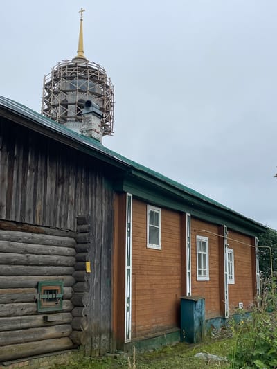 In the city of Кстинино, Russia, a wooden log building is visible in the foreground, its logs weathered and gray. To the right, a section of the building is covered in horizontal wooden planks, painted a warm brown. This section features several white-framed windows with green trim. A green, weathered metal box sits on the foundation near the right side of the brown-planked section.
Behind the buildings, a church dome under scaffolding is prominent. The dome is golden and topped with a cross. The scaffolding is constructed of light-colored wood. A brick chimney protrudes from the roof of the log structure, near the base of the dome. The roof of the log building is covered in dark material, possibly asphalt shingles, and has green trim along the edge. The sky is overcast and a pale gray. Wild vegetation, including tall grasses and flowering plants, grows around the base of the buildings.