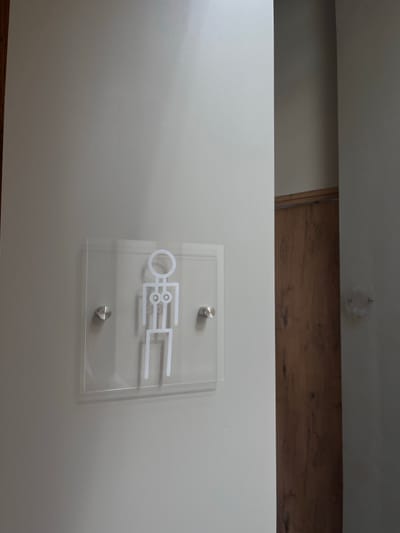 A transparent acrylic sign displaying a stylized white female pictogram is affixed to a light gray wall using two silver stand-off bolts. The pictogram features a circular head, a rectangular torso with two circular representations at the chest, and two rectangular legs. To the right of the sign, a section of a dark brown wooden door is visible, framed by an adjacent darker wall segment. A faint, circular, recessed object or fixture is discernible on the darker wall. The scene is located in Overveen, Netherlands.