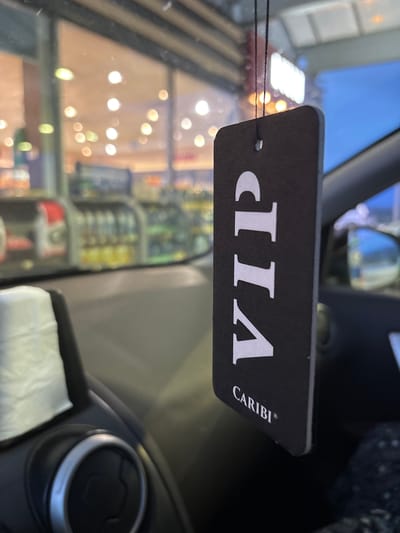 A black car air freshener tag with the word "VIP" in white letters is hanging from the rearview mirror inside a car. The air freshener also has the word "CARIBI" in smaller white letters below "VIP". The background is blurred but appears to be the interior of a convenience store with shelves of products and bright lights, suggesting the car is parked outside. The photo was taken in Kalugerovo, Bulgaria.