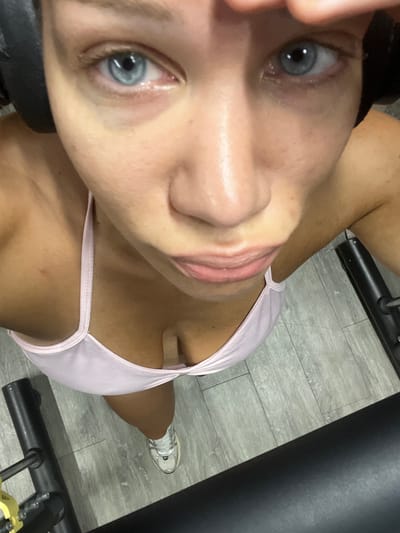 This media file is a close-up, angled photograph taken indoors. The subject is a person with light skin and blue eyes, wearing a light pink sports bra. They have their head tilted back, with their face filling most of the frame. Their gaze is directed upwards, and their lips are slightly parted. The background shows a gray, wood-like floor and parts of exercise equipment, suggesting a gym setting. A single white sneaker is visible on the person's right foot, along with part of their right leg. The lighting appears to be artificial, and there are no discernible weather conditions or time of day cues. No text is visible in the image. The overall impression is a candid moment captured in a fitness environment.