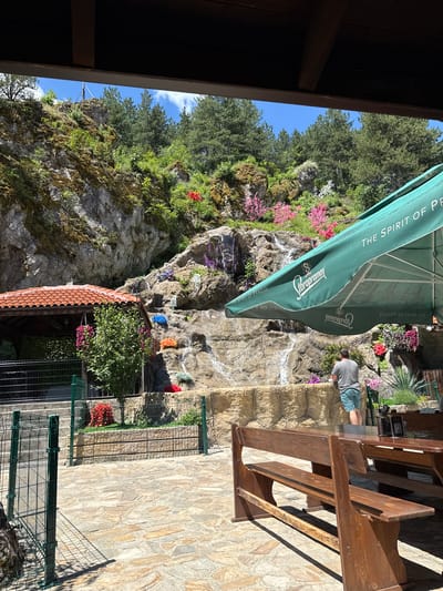 The image depicts an outdoor seating area with a natural rock face backdrop featuring a waterfall. In the foreground, wooden picnic tables and benches are set on a stone patio. A large green umbrella with "Staropramen" branding shades part of the seating area. A man in a grey t-shirt and blue shorts is standing near the waterfall. The rock face is adorned with colorful flowers and greenery, and trees are visible at the top. A small covered structure with a red tile roof is to the left of the scene. The location is identified as Studena, Bulgaria.