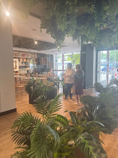 The image depicts the interior of a brightly lit establishment, possibly a cafe or restaurant, with an abundance of artificial plants adorning the ceiling and various planters within the space. The decor features wooden floors, tables, and chairs, creating a warm ambiance.
In the foreground, lush green foliage from potted plants obscures much of the lower portion of the scene. Behind these plants, two women are standing near an open doorway. One woman, wearing a light-colored t-shirt with a graphic and dark pants, is looking to her left. The other woman, dressed in a floral-patterned dress and a scarf, is facing away from the camera.
Further inside, the establishment appears to have a dining area with tables and chairs. Shelving units filled with decorative items are visible against a wall. Through the large glass windows and open doorway, an outdoor area can be seen, with trees and a glimpse of what appears to be a street or plaza. A "SALIDA" (Exit) sign is mounted on a dark column near the doorway. The overall impression is a contemporary interior design focused on incorporating natural elements through extensive plant decoration. The scene is consistent with businesses found in towns like La Roda de Andalucía, Spain, known for their charming and often plant-filled public spaces.