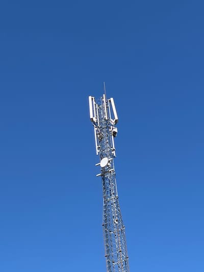 A cell tower is visible against a clear blue sky. The tower is made of metal lattice and has several antennas attached to it. The antennas are white and rectangular. The tower is tall and slender, and it tapers towards the top. The bottom of the tower is not visible in the image. The scene appears to be in an open area, possibly a field or a park, with no other structures or vegetation visible. The location is Kostice, Czechia.