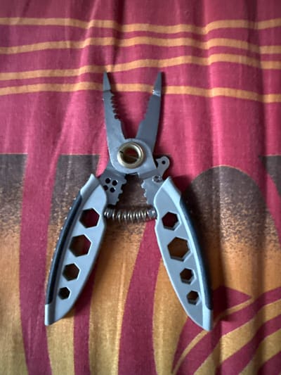 The image shows a pair of gray and silver multi-pliers lying open on a red and yellow striped fabric. The pliers have serrated jaws and a coiled spring in the center. The fabric has a pattern of horizontal stripes, with some darker, blurred shapes visible beneath them. The scene is well-lit and the object is in focus. There are no people or animals visible in the image. The location of Морозовка, Russia is not depicted in the image.
