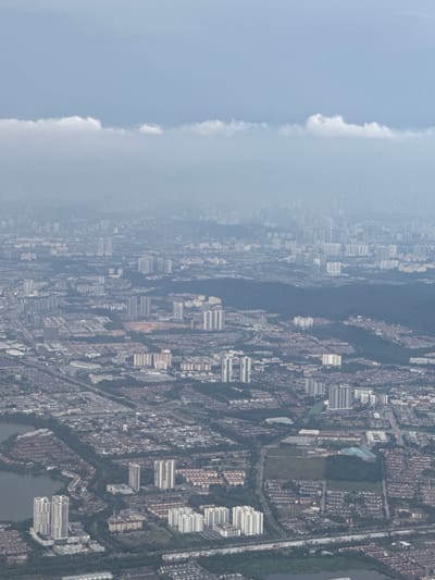 An aerial, medium-shot perspective captures a sprawling urban landscape shrouded in a hazy atmosphere, likely due to pollution or overcast skies. The cityscape extends to the horizon, characterized by a dense concentration of buildings, primarily residential high-rises and apartments. Rows of smaller, terraced houses with red-tiled roofs are also visible, intermingled with the taller structures. Several bodies of water, appearing as dark, reflective surfaces, are present within the urban sprawl, indicating lakes or reservoirs. A network of roads and highways threads through the city, connecting the various neighborhoods. The image exhibits a muted color palette, dominated by shades of grey, blue, and brown, contributing to the overall somber mood. The scene appears to be one of everyday urban life, devoid of any distinct human activity or notable events. The context of the location, identified as Unknown, Malaysia, suggests a tropical or subtropical urban environment.