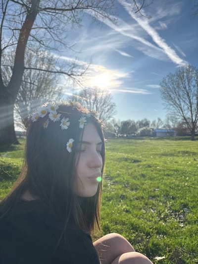 A young person with dark hair lies in a grassy field under a bright sky in what appears to be the city of Beli Brod, Bulgaria. The person is wearing a crown of daisies. The sun is shining brightly, with visible contrails in the sky. There are trees in the background, and grass in the foreground. The scene is peaceful and summery.